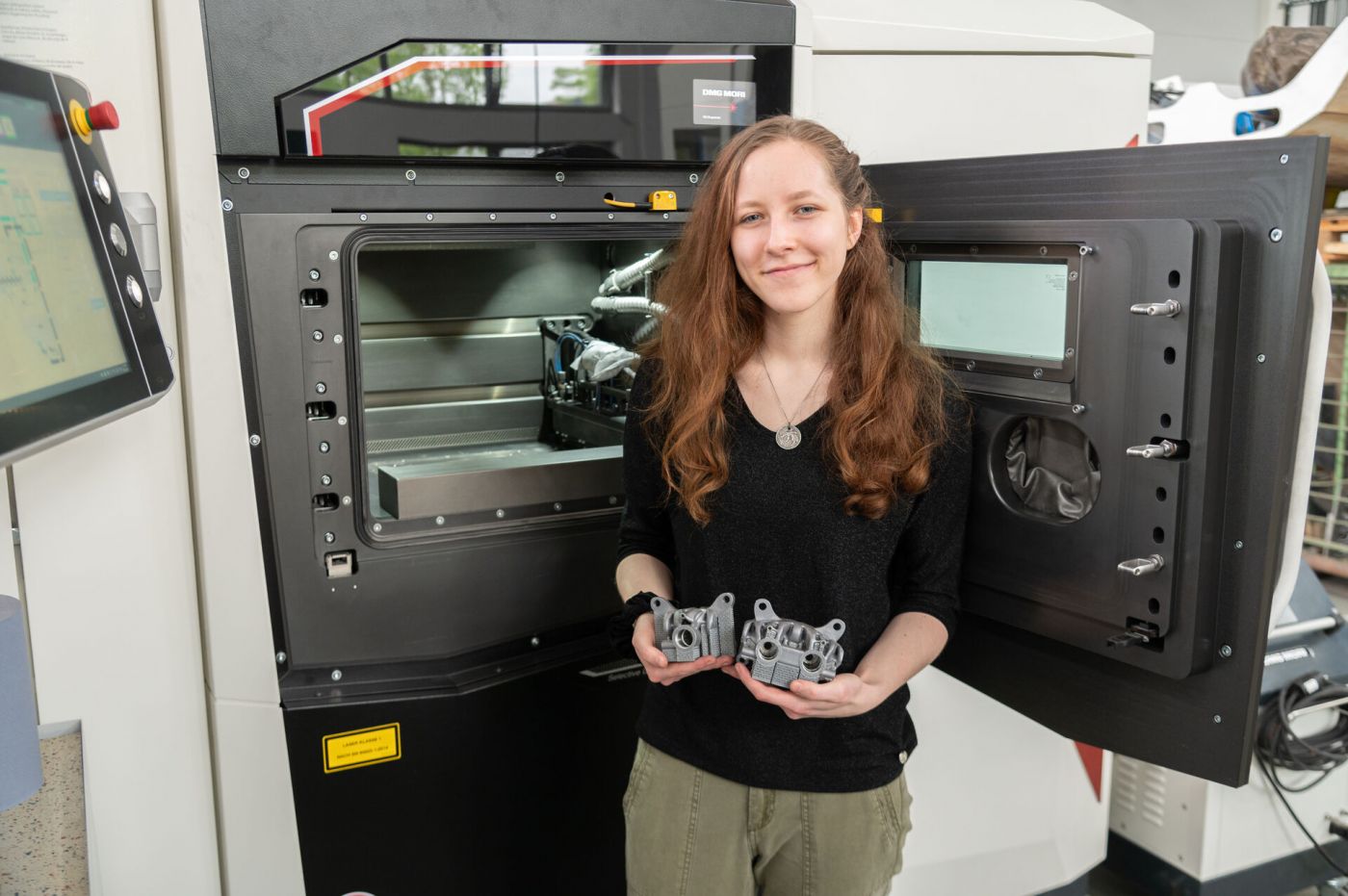 Eine Frau steht vor einem Metall-3D-Drucker und hält zwei Bremssättel in den Händen. (Foto: FH Münster/Frederik Tebbe)