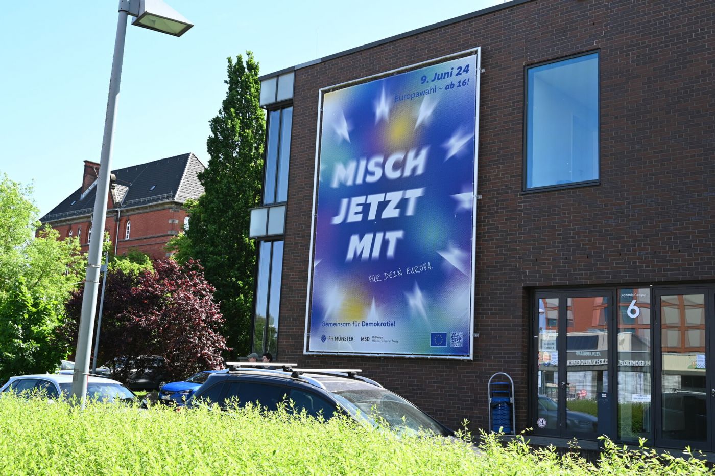 Blaues Banner mit dem Slogan "Misch jetzt mit" an der Fassade eines großen Gebäudes mit Backstein-Glas-Fassade (Foto: Stefanie Gosejohann)