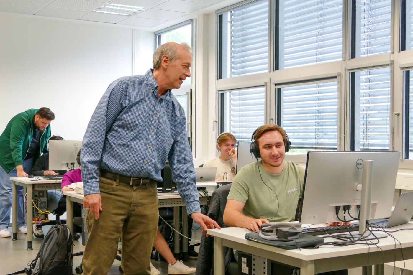 Ein Mann steht neben einem anderen Mann, der am Computer sitzt und Kopfhörer trägt.  (Foto: FH Münster/Jana Bade)
