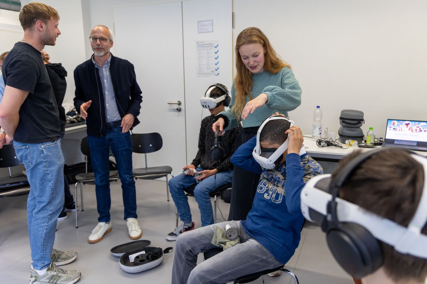 Mehrere Personen sitzen in einem Raum und tragen VR-Brillen, eine Trainerin prüft den korrekten Sitz der Brillen. Daneben stehen zwei Menschen und unterhalten sich. (Foto: FH Münster/Anna Thelen)