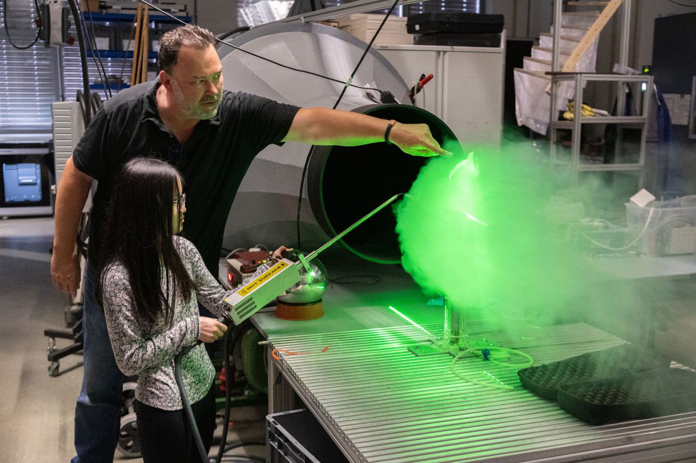 Eine Schülerin erzeugt Nebel an einem Windkanal, der grün leuchtet. Ein Mann hilft ihr dabei. (Foto: FH Münster/Frederik Tebbe)