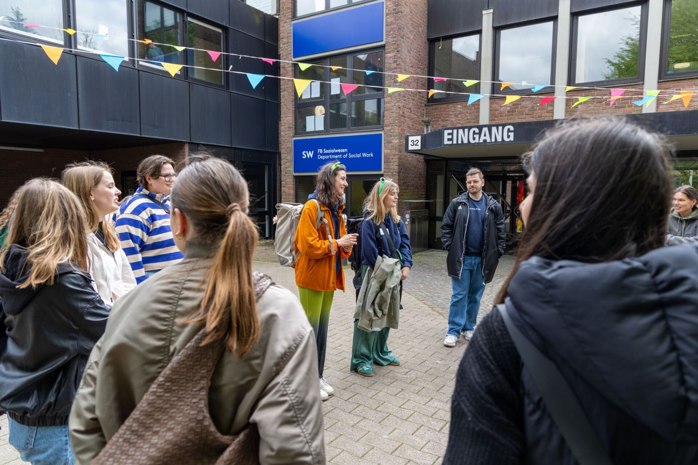 Eine Gruppe von Studierenden steht vor einem Backsteingebäude im Kreis und hört einer Person zu, die spricht. (Foto: FH Münster/Anna Thelen)