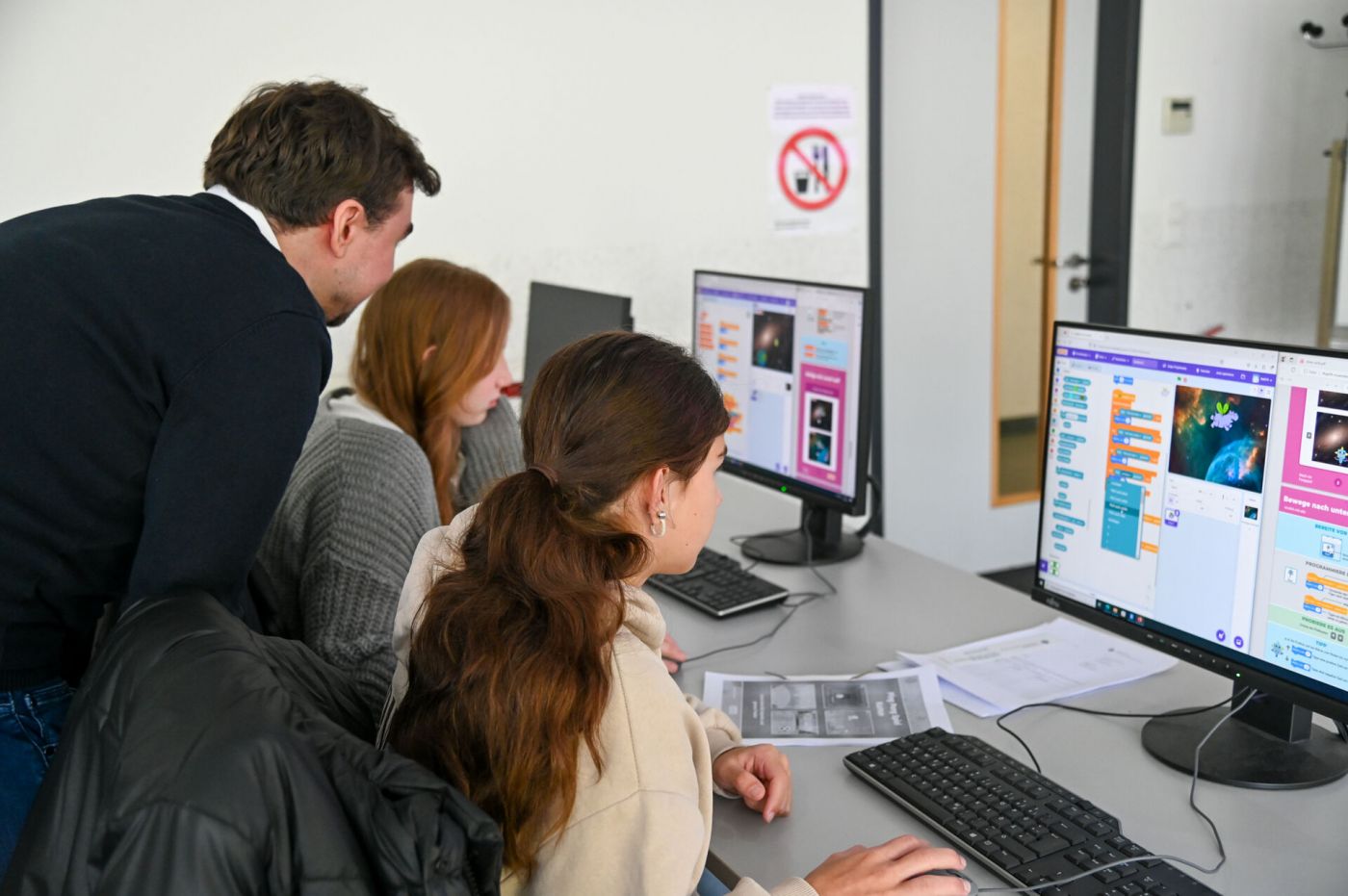 Ein Mann und zwei Schülerinnen blicken auf einen Computerbildschirm.