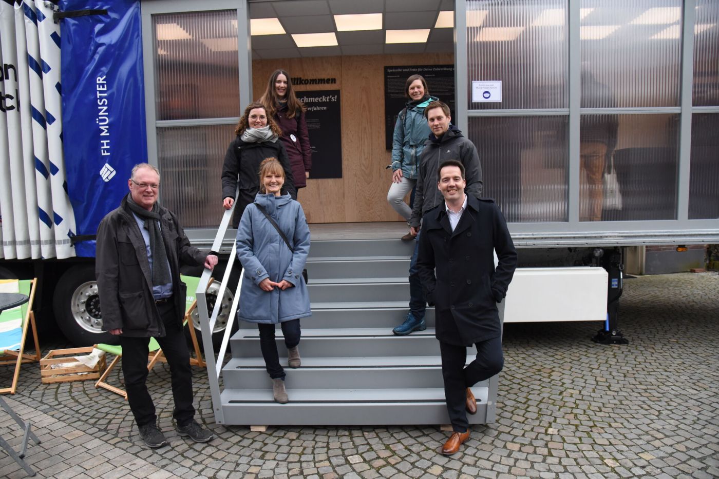 Drei Männer und vier Frauen stehen an der Treppe einer mobilen Ausstellung. (Foto: FH Münster/Frederik Tebbe)