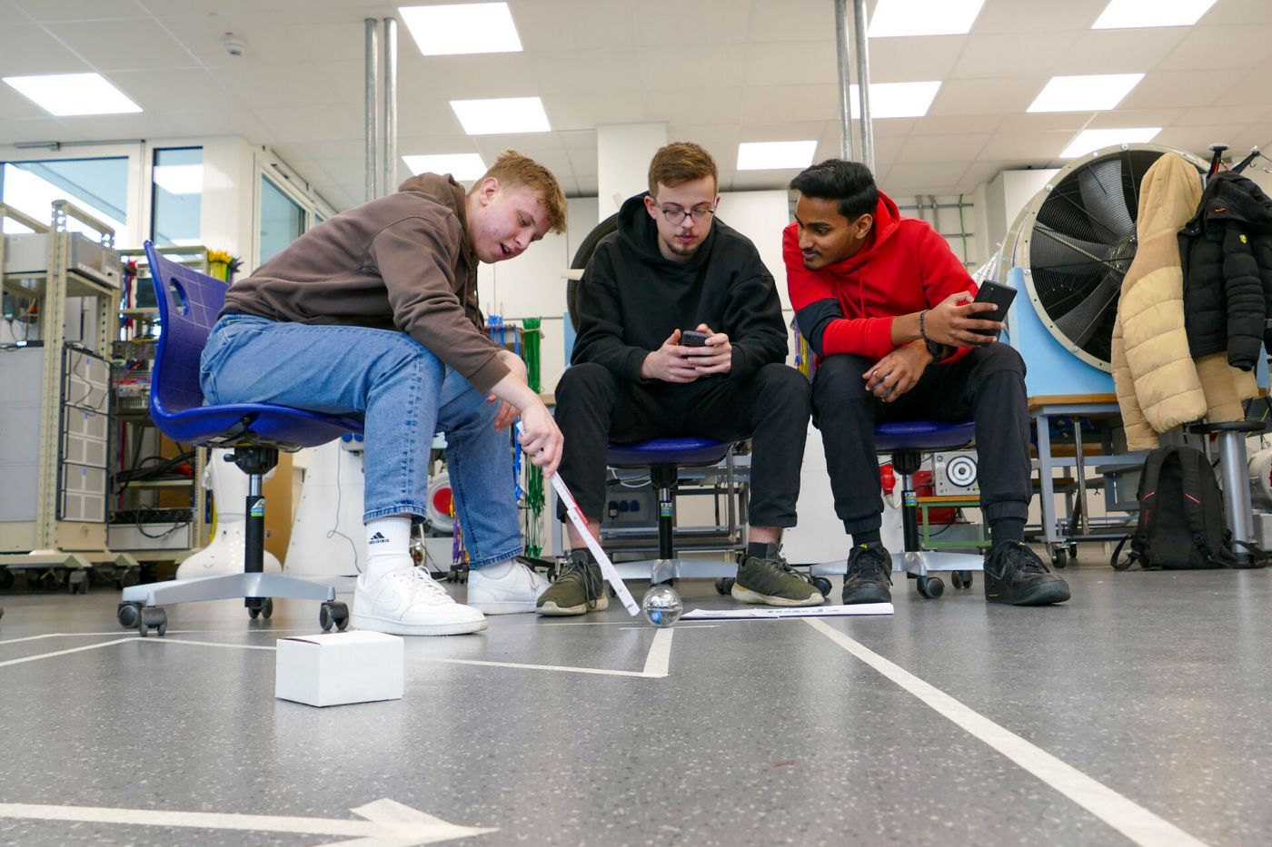 Schülerinnen und Schüler halten ein Smartphone in der Hand und beobachten einen Kugelroboter und einen Pfad aus Klebestreifen auf dem Boden.  (Foto: FH Münster/Jana Bade)