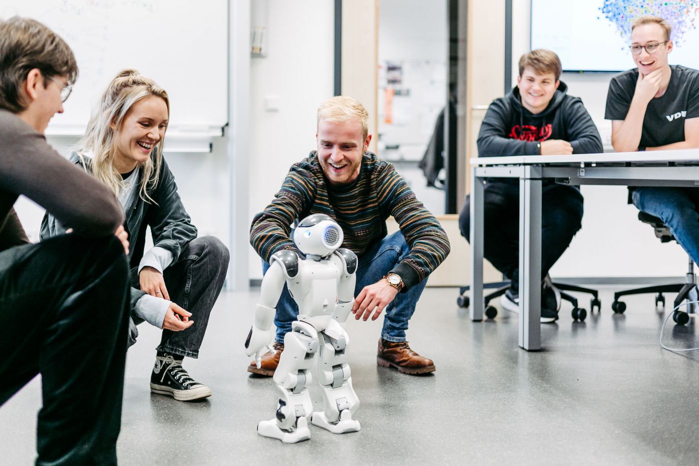 Menschen knien um einen kleinen Roboter, zwei weitere Menschen sitzen an einem Tisch schräg dahinter  (Foto: FH Münster/Anna Haas)