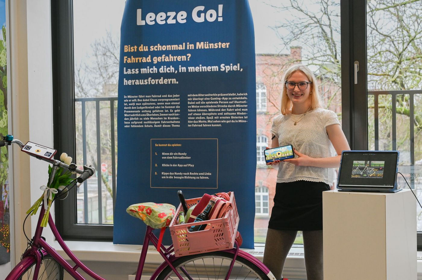 Eine Studentin zeigt eine App auf ihrem Smartphone an einem Ausstellungsstand. Daneben steht ein Fahrrad.