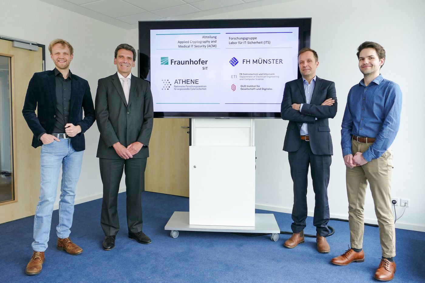 Vier Männer stehen vor einem Bildschirm, der das Logo der FH Münster und eines Fraunhofer-Instituts zeigt. (Foto: FH Münster/Jana Bade)