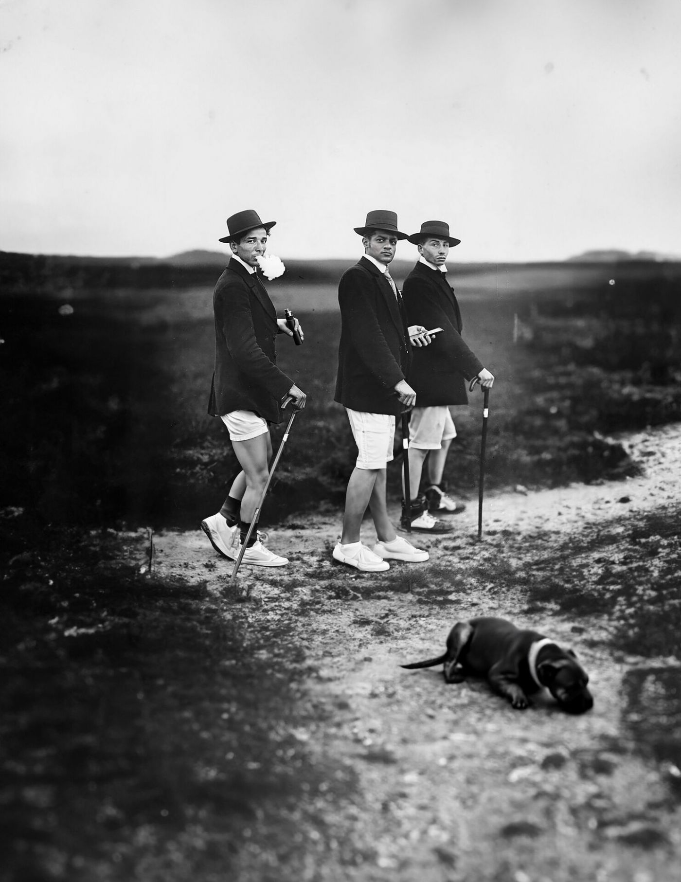 Drei junge Männer, spazieren in hellen Shorts und dunklen Sakkos mit Wanderstöcken in karger Landschaft. Am Wegesrand liegt ein Hund. 