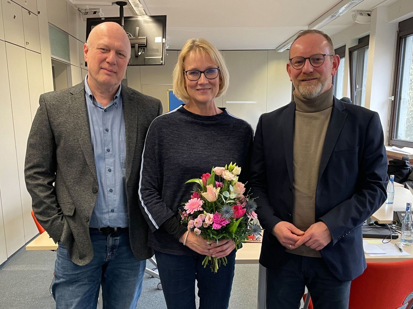 Drei Personen stehen in einem Seminarraum. Die Person in der Mitte hält einen Blumenstrauß in der Hand. (Foto: FH Münster/Anna Thelen)