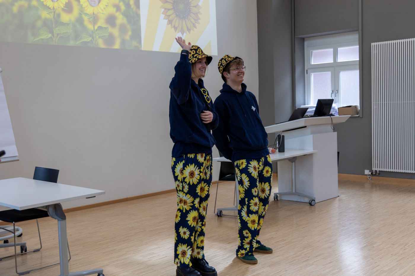 Zwei Personen stehen lächelnd in einem Hörsaal, sie tragen Hüte und Hosen, die mit Sonnenblumen bedruckt sind. Eine der beiden Personen winkt. (Foto: FH Münster/Anna Thelen)