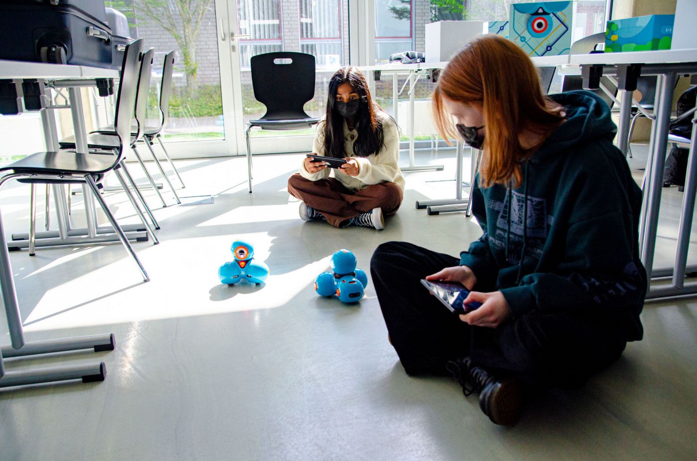 Zwei Mädchen sitzen auf dem Boden und steuern zwei kleine Roboter, die umherfahren. (Foto: FH Münster/Frederik Tebbe)