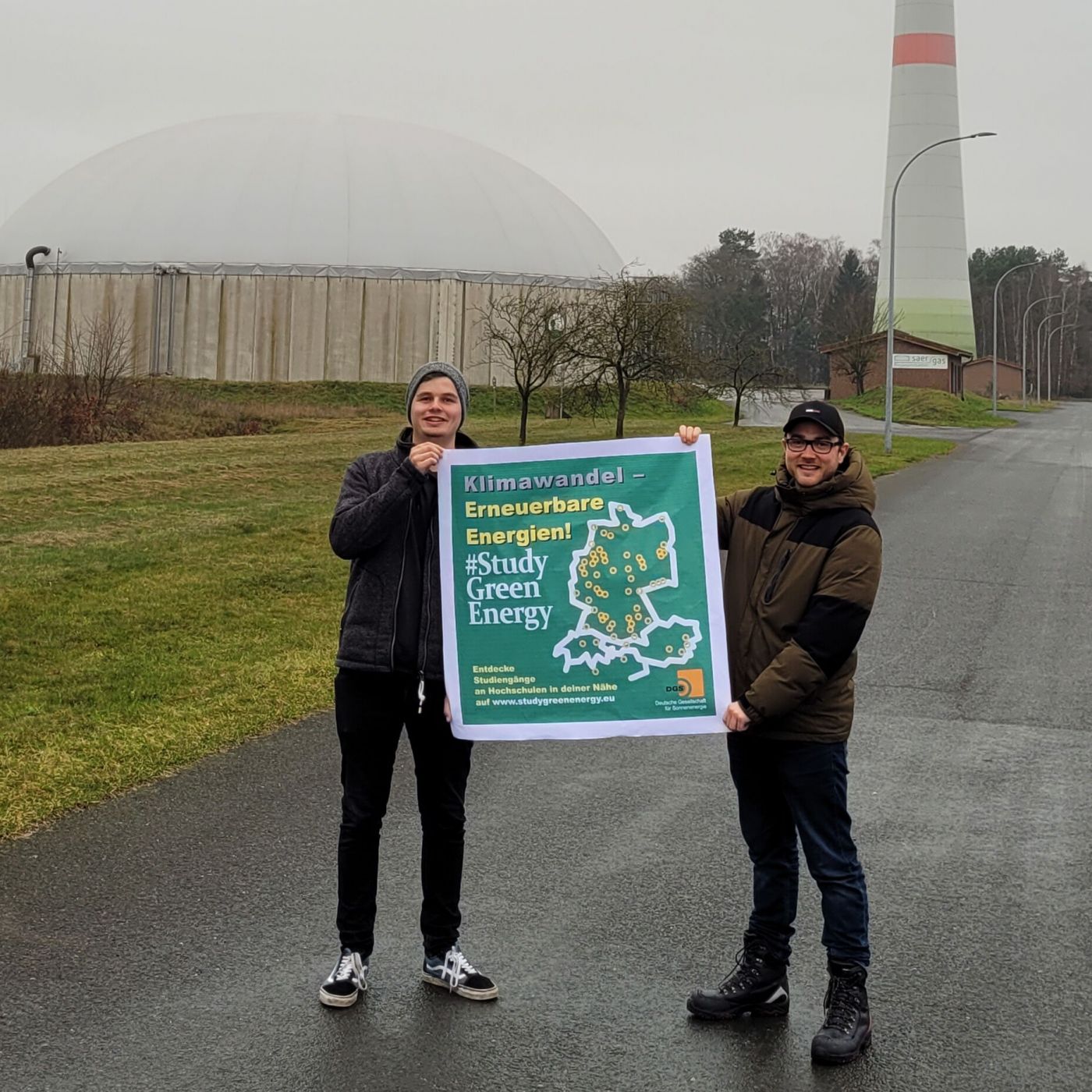 Zwei Studenten halten ein Plakat hoch. Im HIntergrund ist eine Windkraftanlage zu sehen. (Foto: FH Münster/FB EGU)