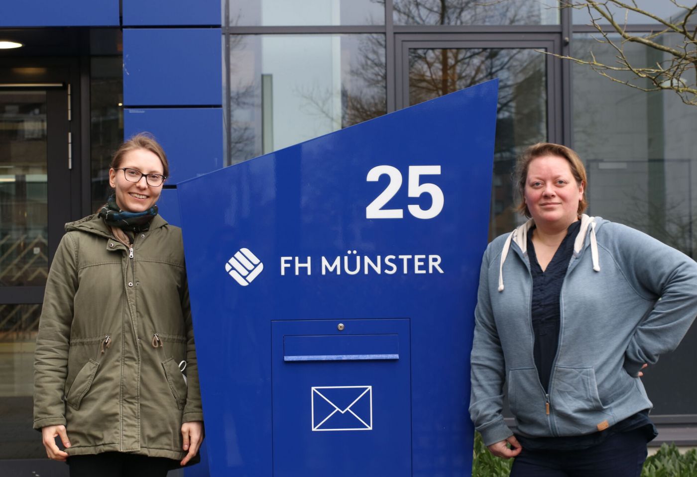 Zwei Frauen neben dem Logo der FH Münster.