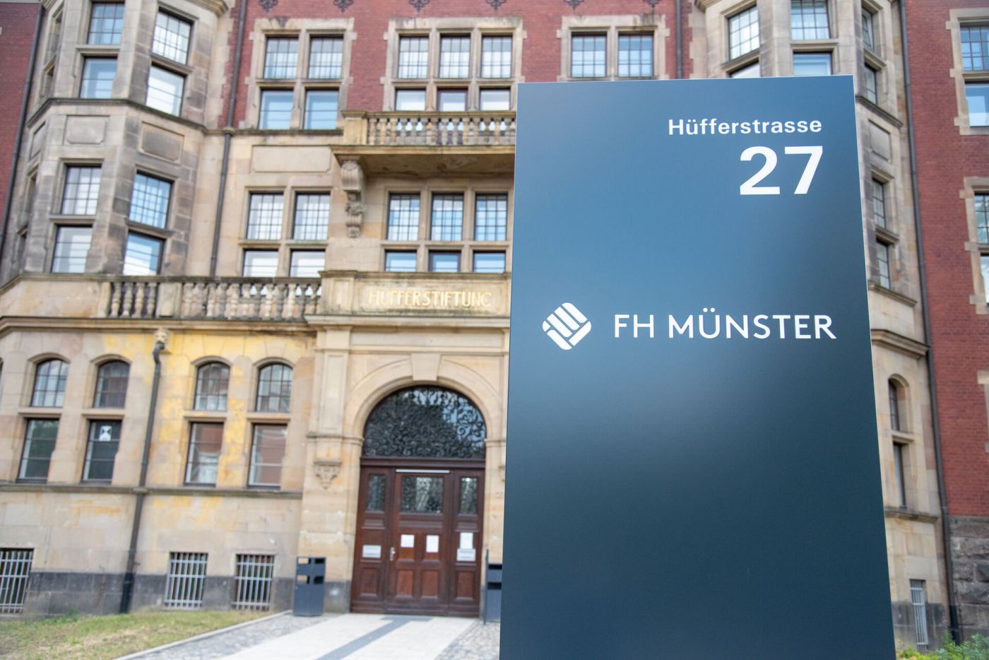 Der Haupteingang der Hüfferstiftung ist im Hintergrund zu sehen - vordergründig ist eine Stele mit der Adresse - Hüfferstrasse 27 - sowie dem Logo der FH Münster zu sehen.