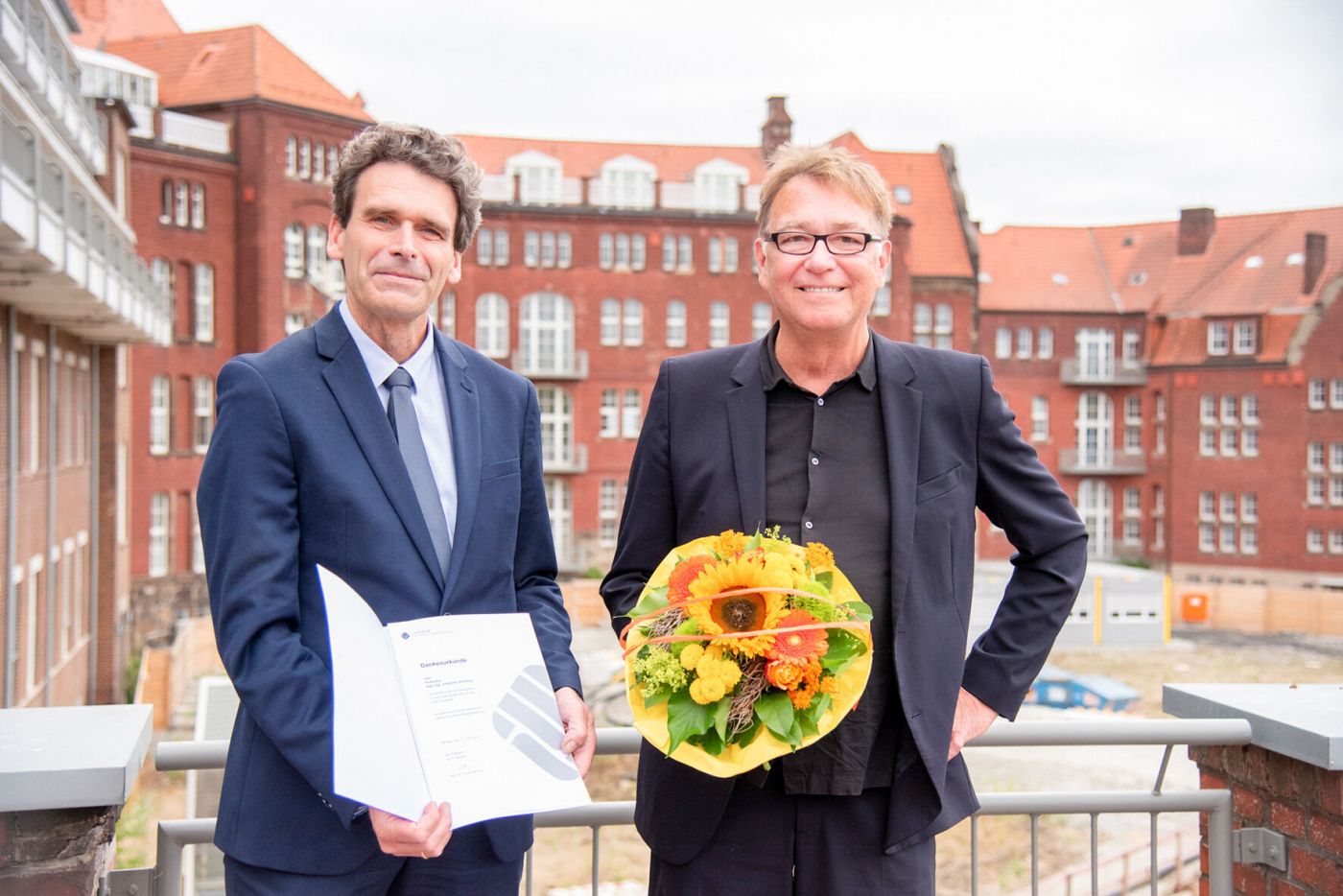 Zwei Männer stehen auf einer Terrasse mit einem Blumenstrauß und einer Urkunde. (Foto: FH Münster/Michelle Liedtke)