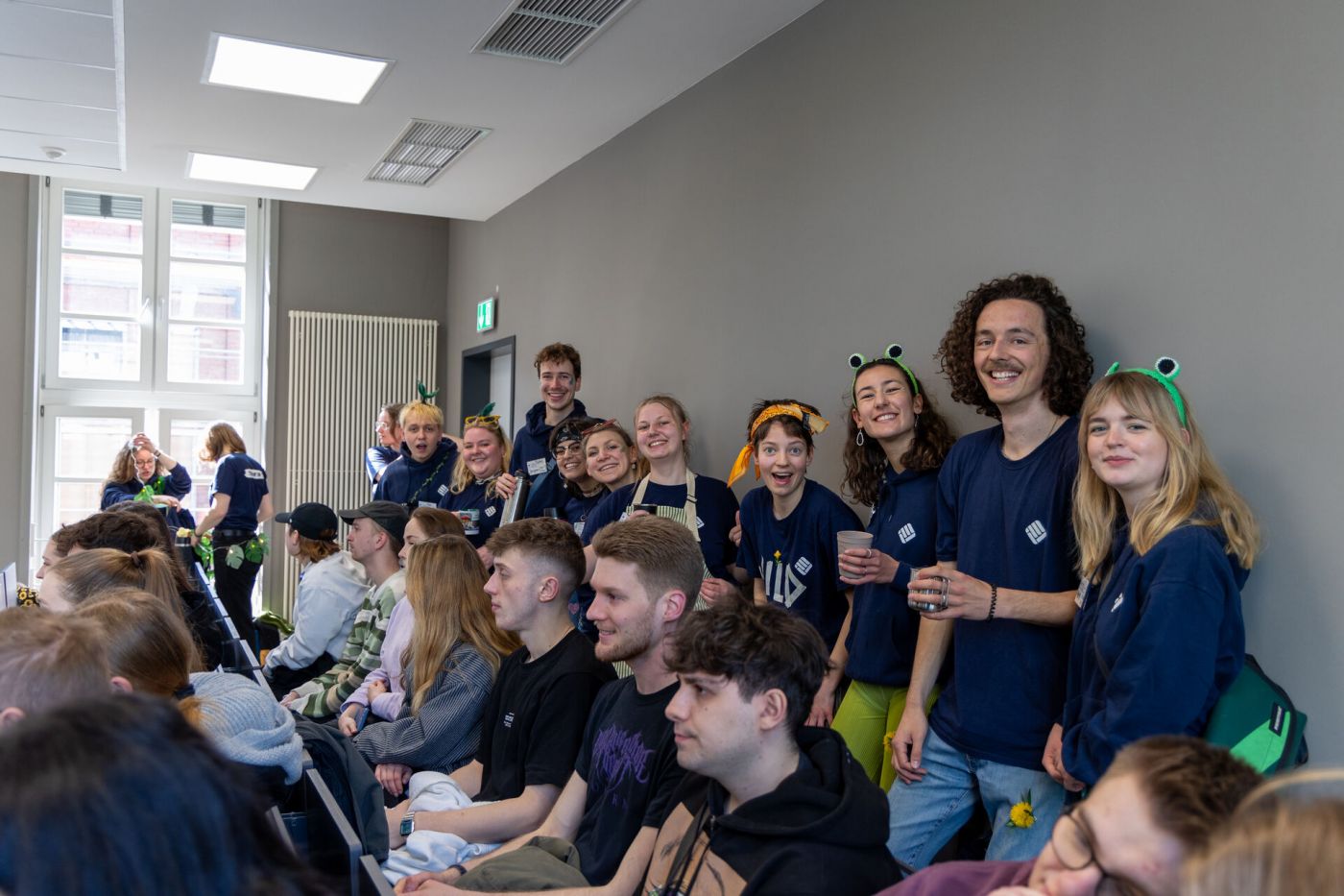 Eine Gruppe junger Menschen steht in einem Hörsaal und lächelt in die Kamera, sie tragen einheitliche blaue Pullover und dazu unterschiedliche Verkleidungen. (Foto: FH Münster/Anna Thelen)