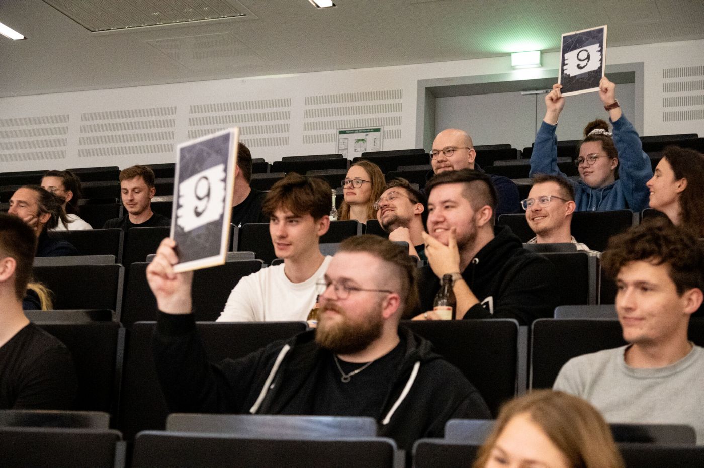 Studierende im Publikum halten Punktetafeln mit dem Wert neun hoch. (Foto: FH Münster/Frederik Tebbe)