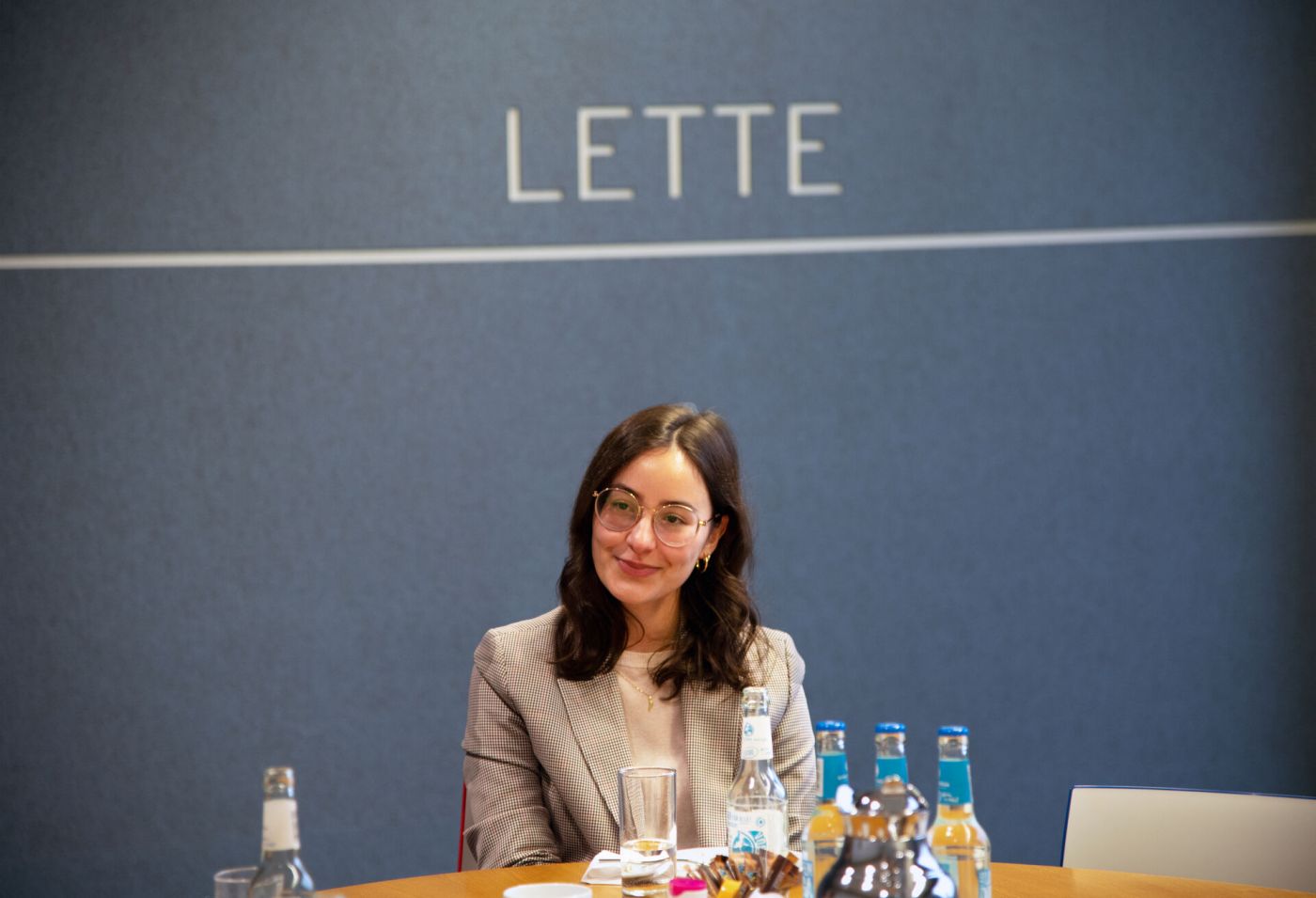 Eine junge Frau sitzt an einem Tisch. Hinter ihr ist eine blaue Wand mit der Aufschrift "Lette" zu sehen.  