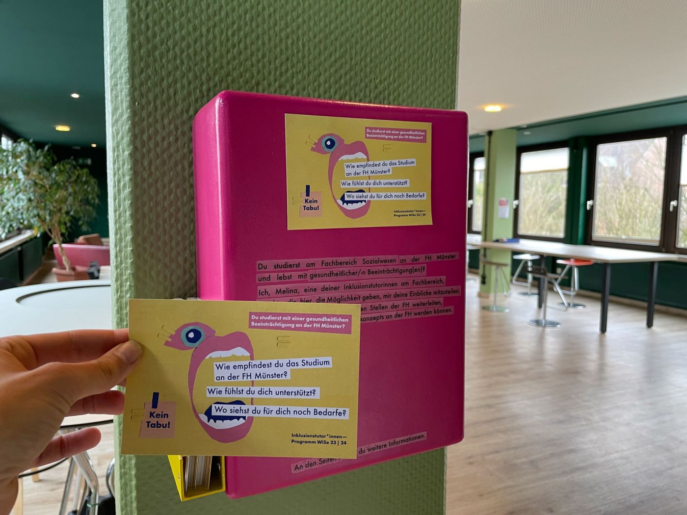 Eine Hand hält eine gelbe Postkarte, im Hintergrund hängt ein pinker Briefkasten. (Foto: FH Münster/Anna Thelen)