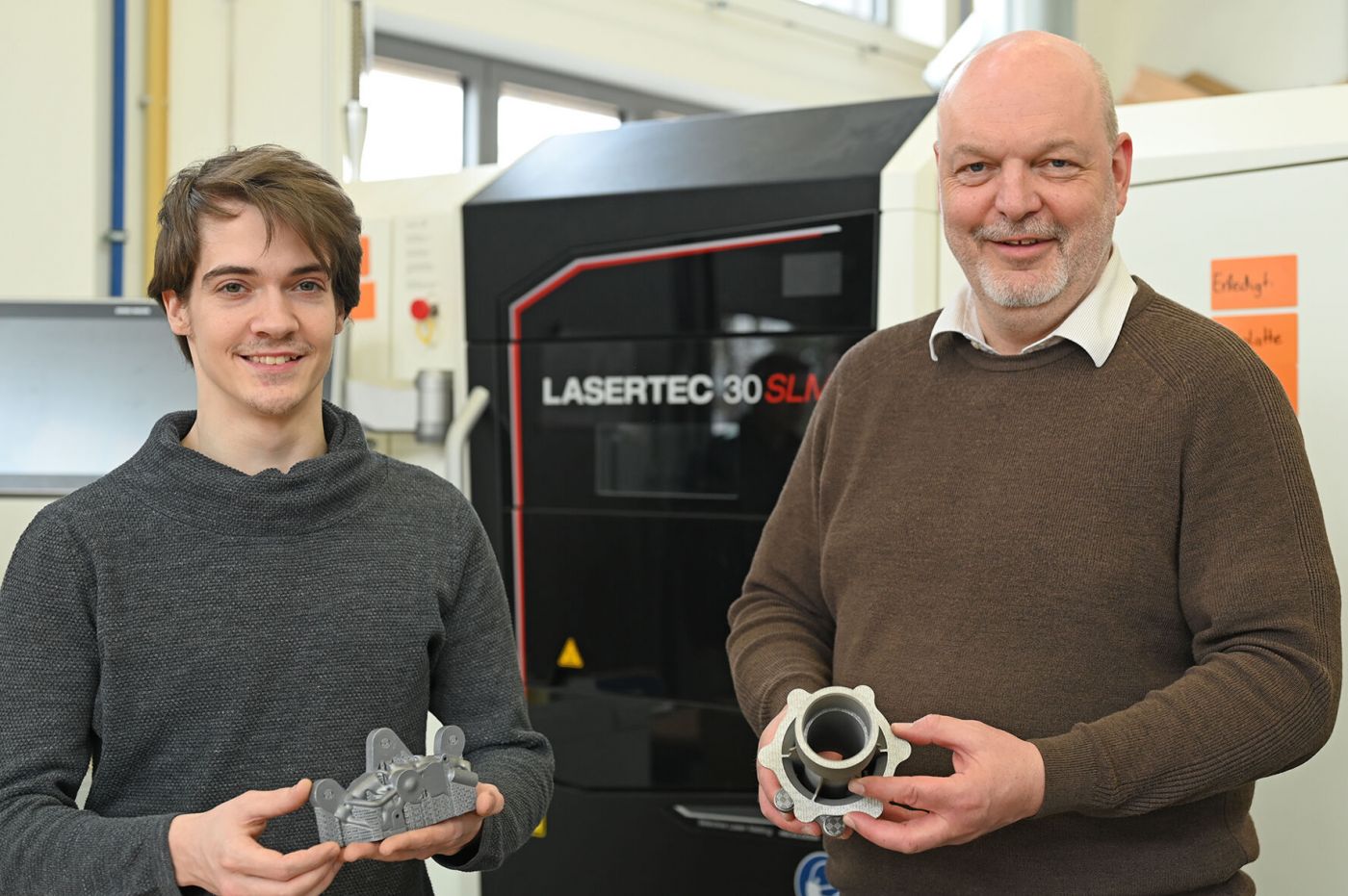Zwei Männer vor 3D-Metalldrucker halten Metallbauteile in den Händen, die mit dem Drucker gefertigt wurden. (Foto: Stefanie Gosejohann)