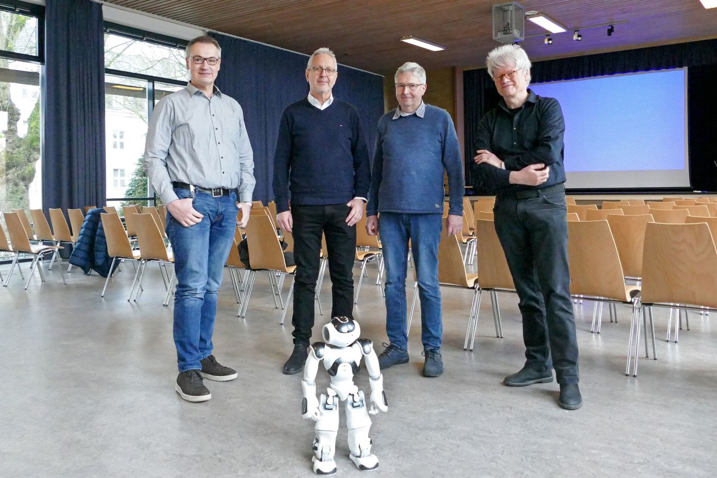 Vier Männer und ein kleiner Roboter stehen in einer Schulaula. 