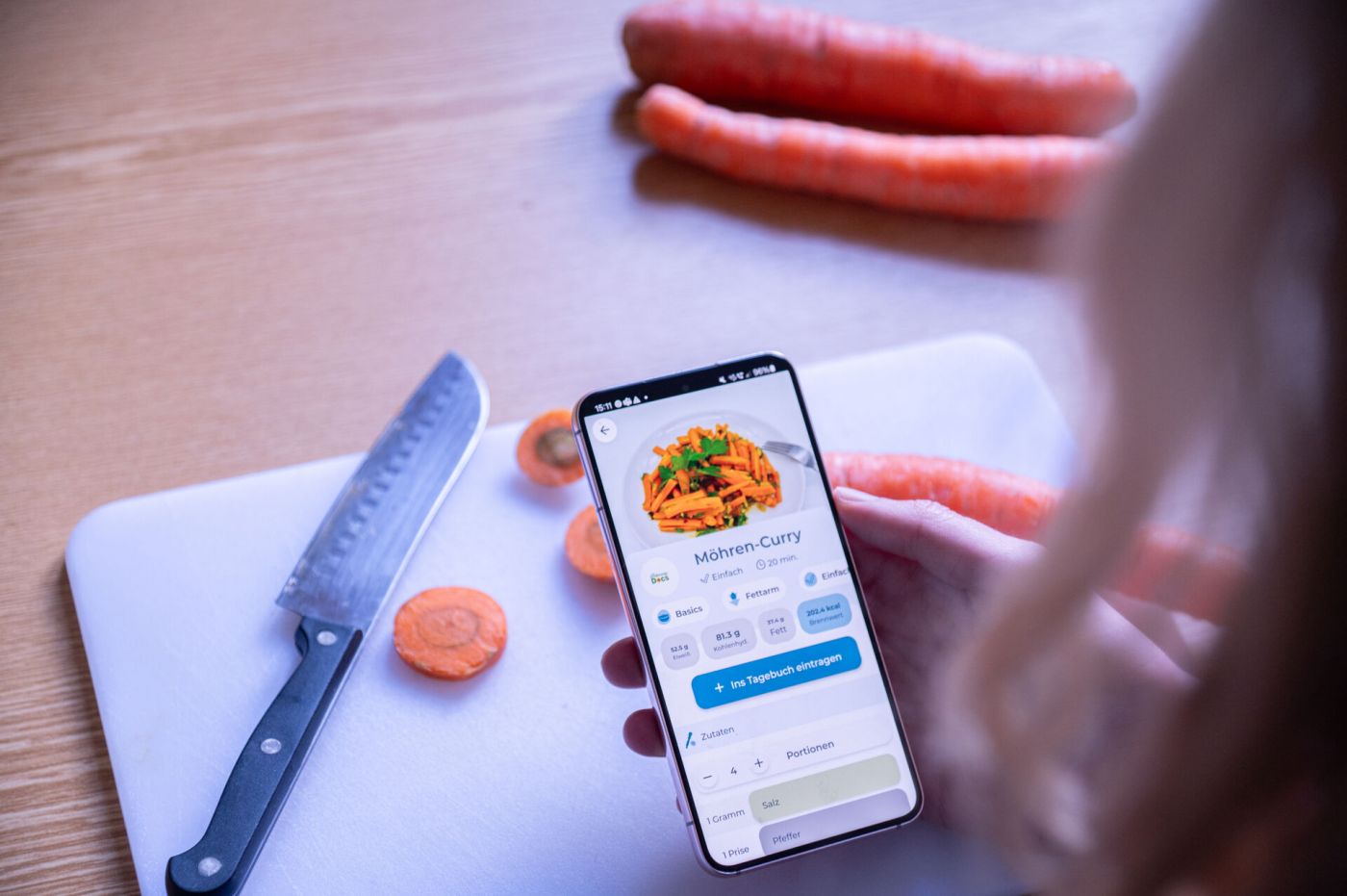 Eine Frau blickt auf ein Smartphone, auf dem die App meal&heal läuft. Im Hintergrund ist ein Schneidebrett samt Messer zu sehen, auf dem eine Möhre geschnitten wurde. (Foto: FH Münster/Frederik Tebbe)