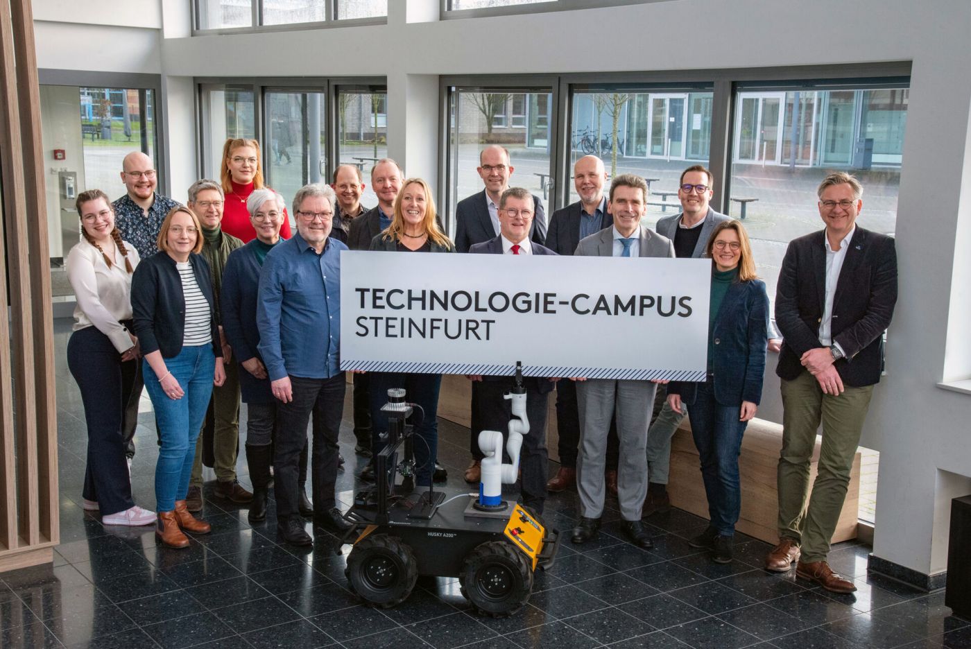 Ein Gruppenfoto auf dem Steinfurter Campus der FH Münster, bei dem ein Roboterarm ein Schild mit der Aufschrift 