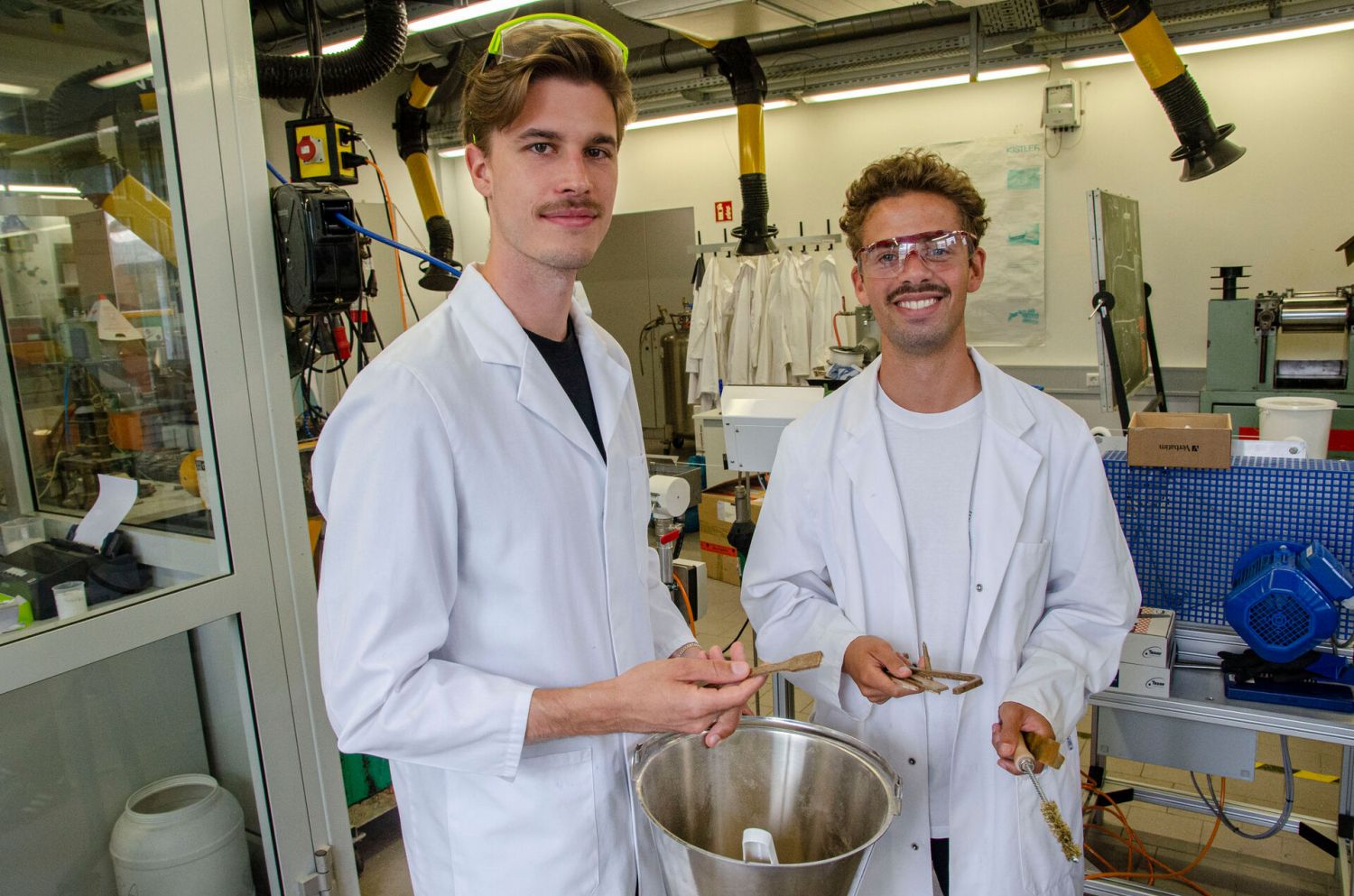 Zwei Männer stehen mit Kittel und Schutzbrille in einem Labor.  Bence Ridder (l.) und Calvin Middel untersuchen für ihre Bachelorarbeit am Fachbereich Design, wie man aus dem Reststoff von Zuckerrohr recycelbare Alltagsgegenstände herstellen kann.