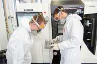 Zwei Männer in Laborkitteln begutachten eine Probe aus einem Metall-3D-Drucker.