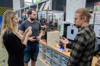 Zwei Männer und eine Frau unterhalten sich in einer Werkstatt und begutachten Teile aus dem 3D-Drucker.